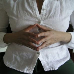 woman in white shirt, sitting with her fingertips lightly pressed together